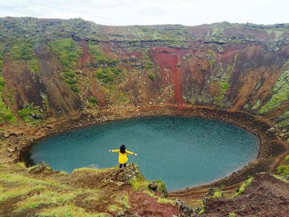 The crater of Kerið, it's aquamarine water and red flanks and a lady with a yellow jacket in front of it.