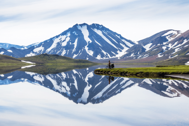 Montañas nevadas en las Tierras Altas de Islandia reflejadas en un lago con una persona en bicicleta al frente.