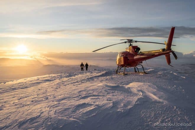 deux personnes se tenant la main au sommet de l'une des montagnes entourant Reykjavik à côté de l'hélicoptère qui les a amenés là-bas