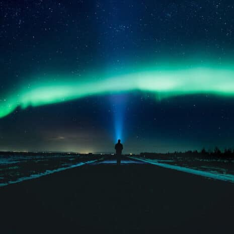 Une personne debout dans une rue sous les aurores boréales, braquant une lampe de poche dans le ciel.
