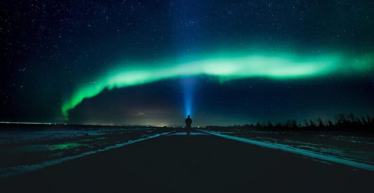 A person standing in a in a street under the northern lights, shining a flashlight into the sky.