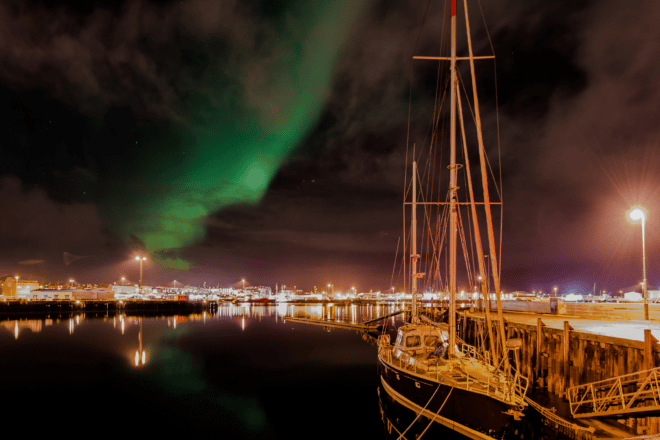A sailboat in the foreground with northern lights dancing over Reykjavik Old Harbour