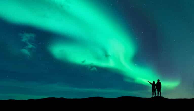 A silhouette of a couple standing under green northern lights.