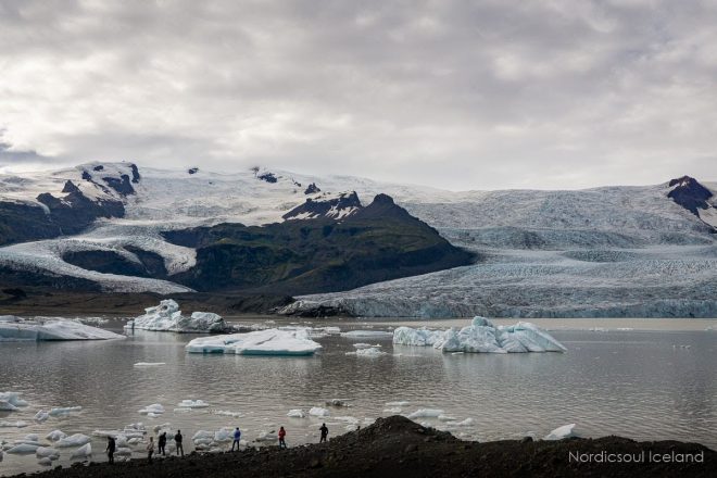 Personas de pie en la costa de una laguna glaciar frente a una enorme capa de hielo