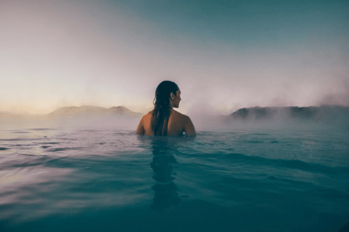 Una mujer desde atrás sumergiéndose en la Laguna Azul de Islandia, con poca luz y vapor saliendo del agua.