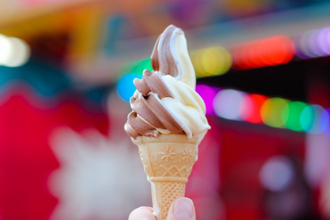 Hand holding chocolate and vanilla soft ice cream in waffle cone with blurred bright colorful amusement park at background.
