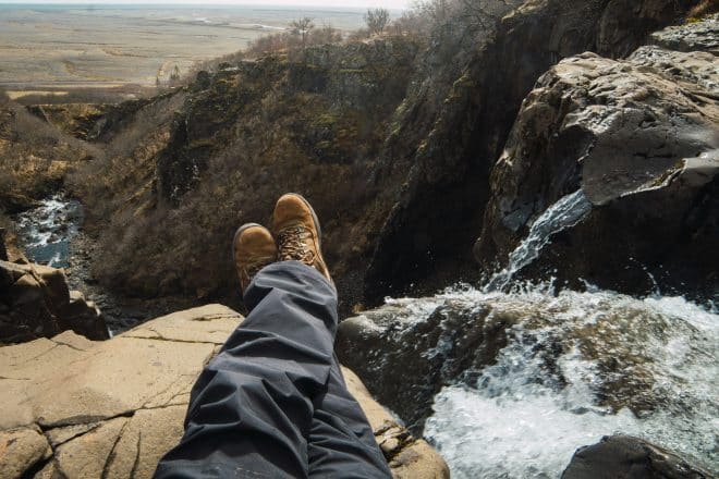 Recortar las piernas del hombre sentado en la cima de la montaña cerca de la cascada en ropa de invierno