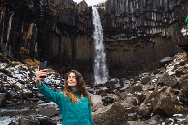 Una mujer parada frente a la cascada Svartifoss en Islandia, tomándose una selfie en un teléfono inteligente