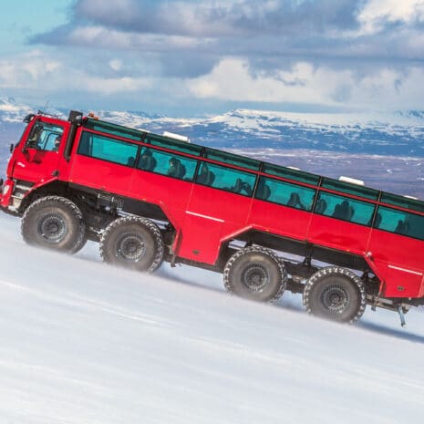 Un gran camión rojo subiendo por un glaciar en Islandia con montañas al fondo.