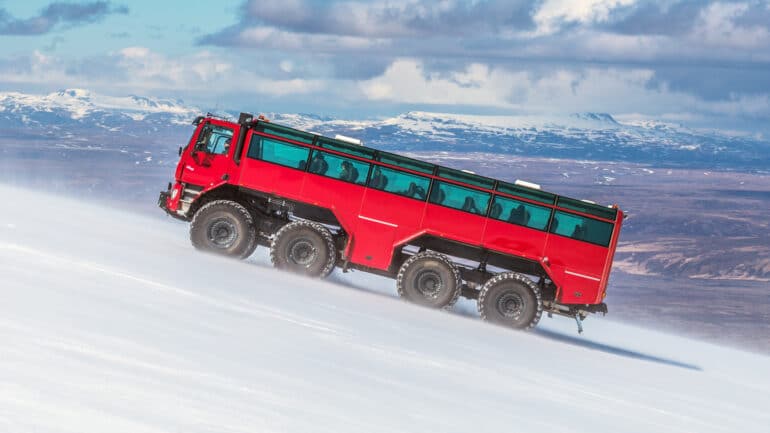 A large red truck driving up a glacier in Iceland with mountains in the background.