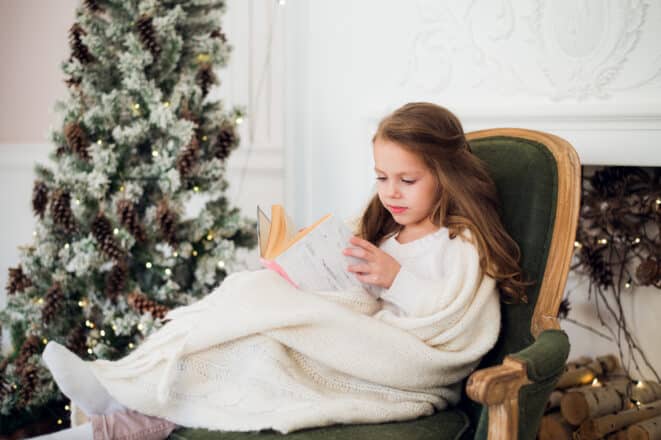 A little girl reading a book near a Christmas tree
