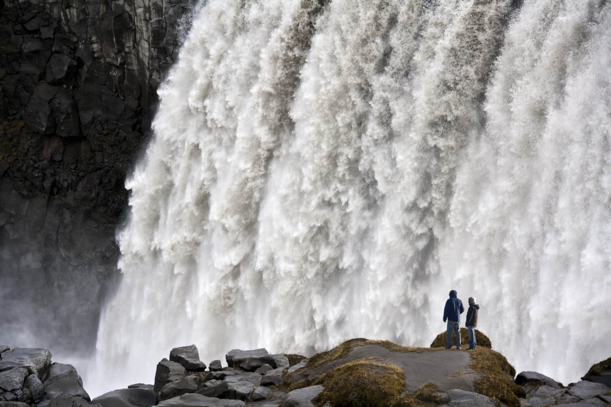 Dos personas de pie frente a la enorme cascada Dettifoss en el norte de Islandia.