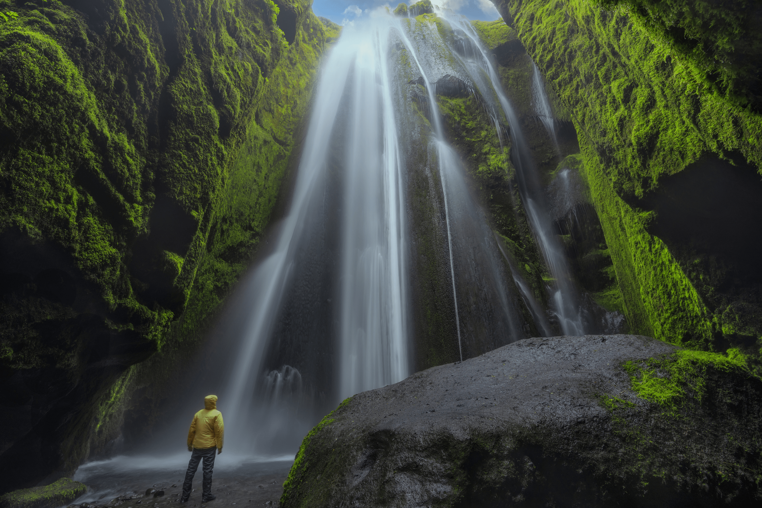 A man standing inside of a moss-covered cave in front of Gljúfrabúi Waterfall in South Iceland