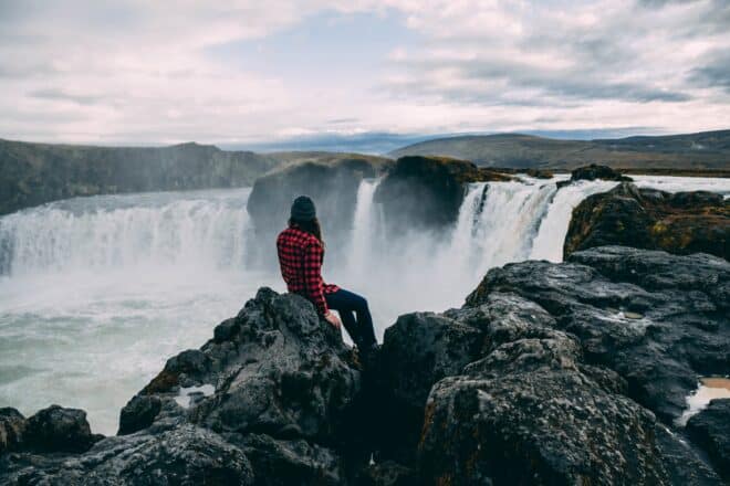 Una mujer sentada sobre las rocas frente a la cascada de Goðafoss en el norte de Islandia.