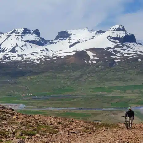 Snow-topped mountains of East Iceland in the back with a man and a mountain bike on a gravel path in front.