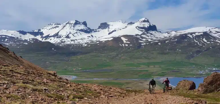 Snow-topped mountains of East Iceland in the back with a man and a mountain bike on a gravel path in front.