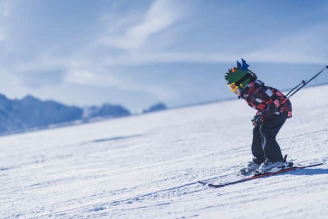 Child skiing in mountains. Active teenage kid with safety helmet, goggles and ski poles running down ski slope.  Snowy landscape, sunny day in winter season.