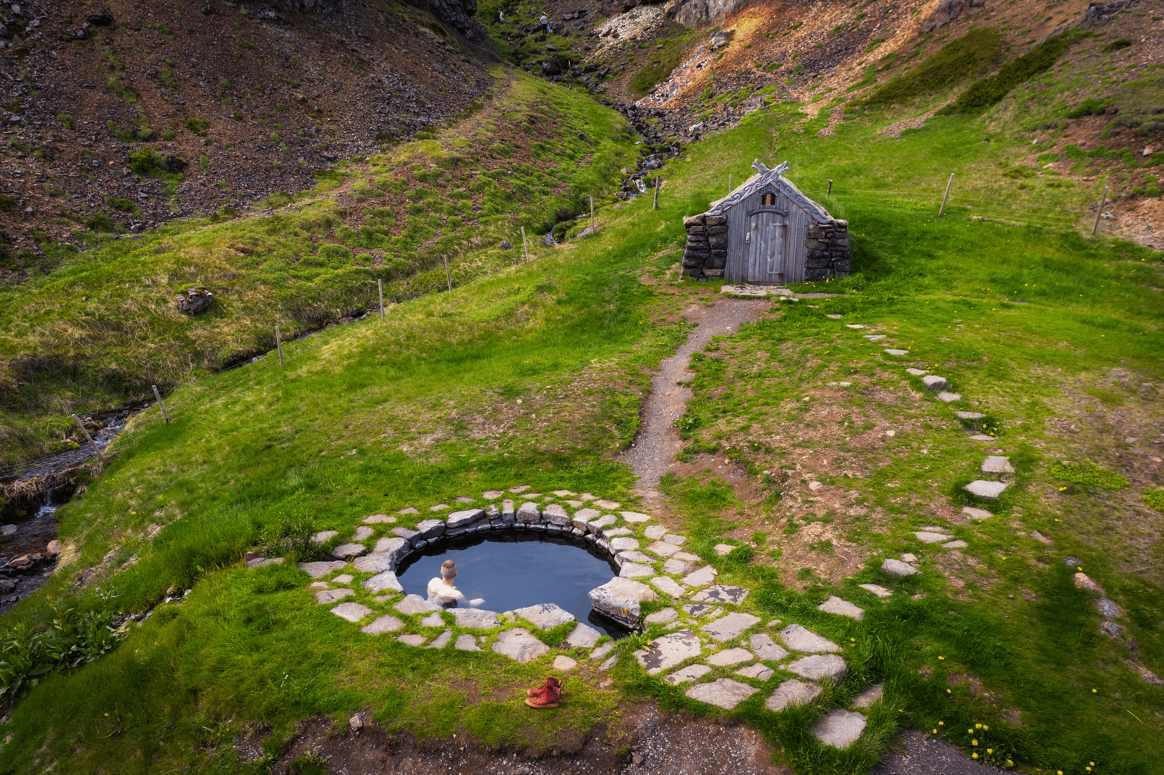 A woman relaxing in Gudrunarlaug Pool in West Iceland, a small turh house nearby.