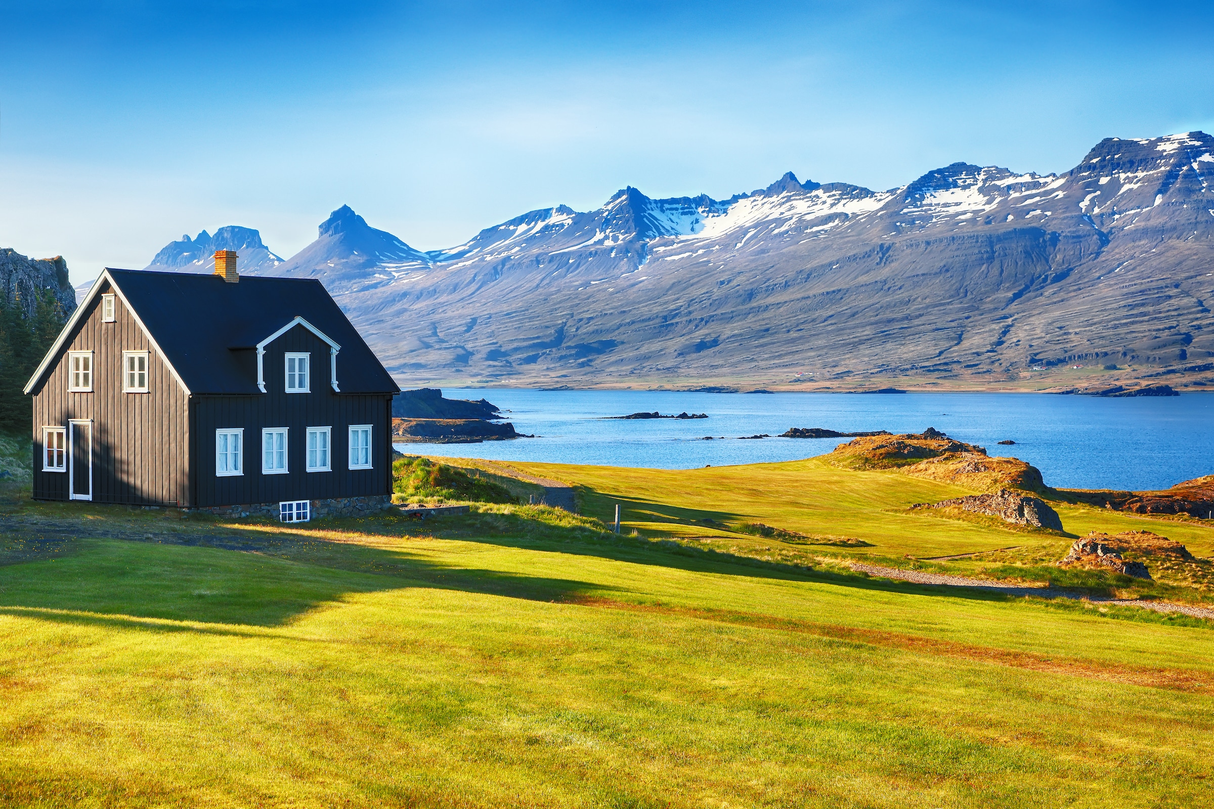 Una antigua Casa Negra junto al océano frente a la hierba verde con montañas al fondo en el este de Islandia.