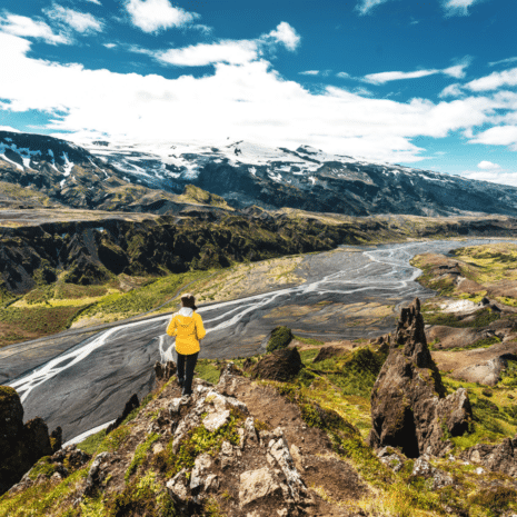 A person standing on a cliff, looking at snow-capped mountains, black sands and a river in Thorsmork Valley, Iceland.