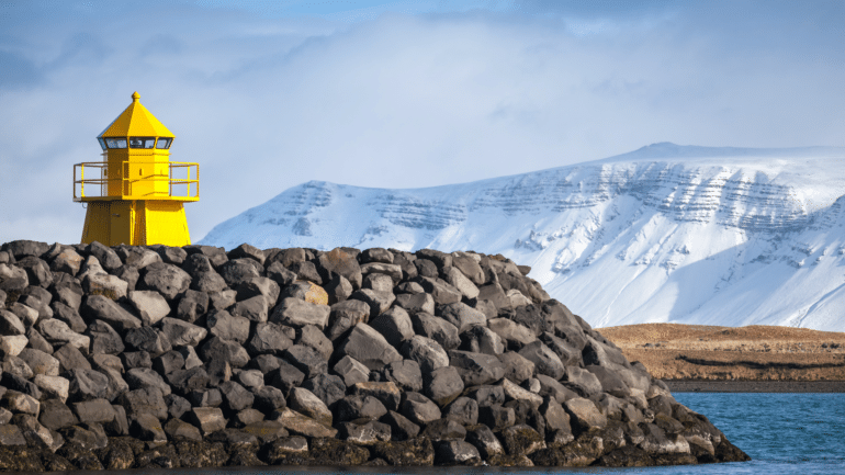 Un faro amarillo en Reykjavik con el monte Esja cubierto de nieve al fondo.