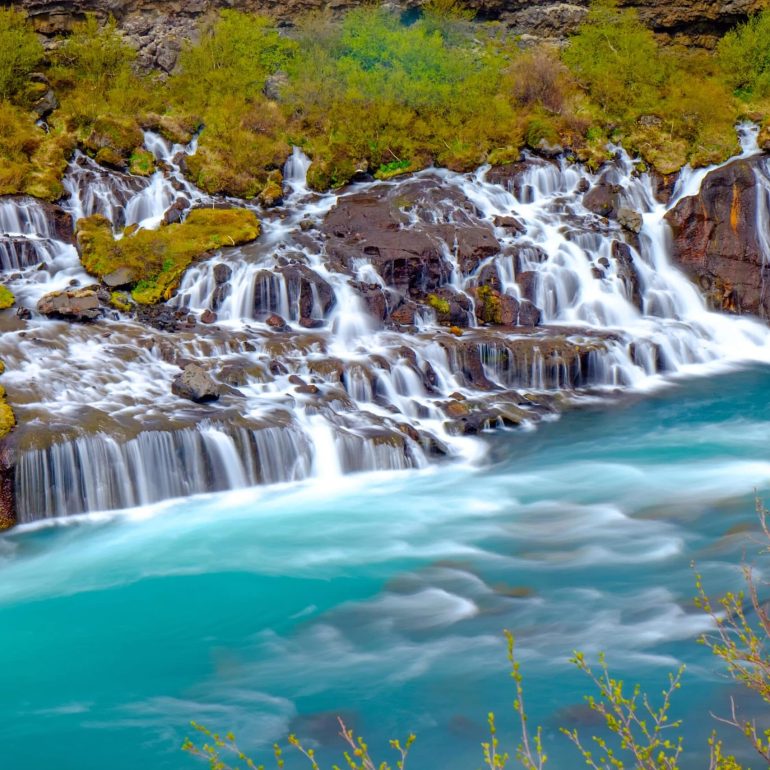 Water flowing from a lava field and into a blue river, forming Hraunfossar Waterfall in Iceland.